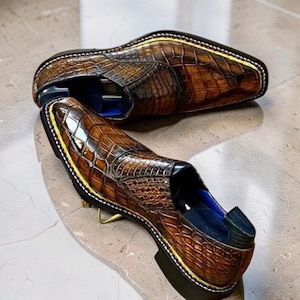 May include: A pair of brown leather loafers featuring a textured crocodile skin pattern with a glossy finish. The shoes have a pointed toe, a dark sole with a prominent yellow welt stitching, and a deep blue interior lining visible at the heel.