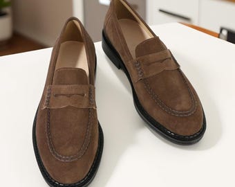May include: A pair of brown suede loafers featuring a classic penny slot strap across the vamp, visible stitching along the rounded toe, a smooth beige interior lining, and a solid black rubber sole with a textured edge detail.