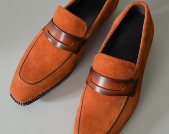 Könnte beinhalten: Ein Paar Loafer aus orangefarbenem Wildleder mit einem glatten, dunkelbraunen Lederriemen über dem Vorderblatt. Die Schuhe haben eine abgerundete Zehenpartie, einen sichtbaren genähten Rahmen entlang der Sohle und ein schwarzes Innenfutter.