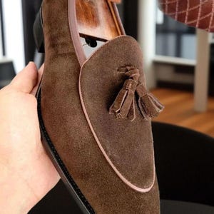 Puede incluir: Un mocas&iacute;n de ante marr&oacute;n con una borla decorativa en el empeine, un ribete de cuero liso a lo largo del cuello y una horma de madera insertada en el interior. El zapato tiene una suela negra y una construcci&oacute;n de vira cosida visible.