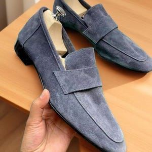 May include: A pair of slate blue suede loafers featuring a classic apron toe design, a wide strap across the vamp, and a low black heel. Each shoe is fitted with a wooden shoe tree that includes a polished silver metal handle at the top.