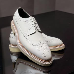 以下が含まれることがあります： 装飾的な穴あきと明るい色のソールを備えた、白い革製のブローグシューズのペア。靴はクラシックなレースアップデザインで、革のアッパーは質感があります。ソールには明るい茶色のトリムが付いています。