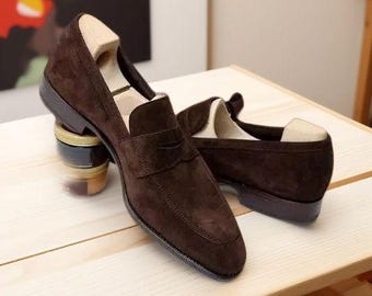 May include: A pair of dark brown suede penny loafers featuring a classic moccasin-style toe construction, a horizontal strap across the vamp, and a low stacked heel. Each shoe contains a light-colored wooden shoe tree insert for shape retention.