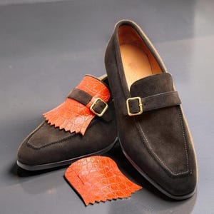 May include: A pair of dark brown suede loafers featuring a gold-toned buckle strap. One shoe includes a detachable bright orange textured fringe overlay, and a matching loose orange fringe piece is placed in front of the shoes.
