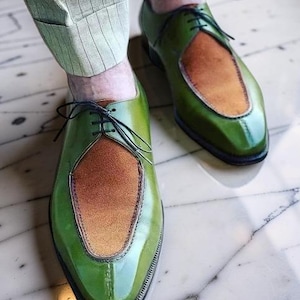 May include: Green and brown leather dress shoes with black laces. The shoes have a glossy finish and a unique design with a brown suede insert. The shoes are paired with light green pinstripe pants.