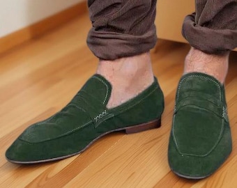 May include: A pair of dark green suede loafers featuring a classic penny slot design across the vamp, visible white contrast stitching along the seams, and a low-profile brown heel, worn with brown cuffed trousers.