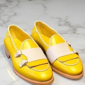 May include: A pair of bright yellow leather loafers featuring a wide off-white strap across the vamp. Each shoe is secured with two polished silver-tone metal buckles on the side and sits on a light tan wooden sole with a low heel profile.