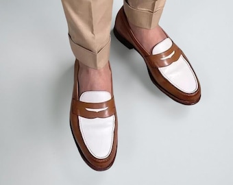 May include: A pair of two-tone penny loafers featuring a tan leather body with a white leather vamp and a classic strap across the top. The shoes are worn with light tan cuffed trousers, showcasing a sleek design with visible stitching details.