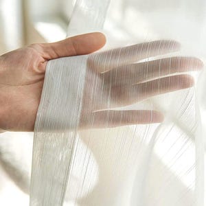 Könnte beinhalten: Ein transparenter, weißer Vorhang mit einer zarten, vertikal gestreiften Textur. Der Stoff ist lichtdurchlässig und weist ein feines, lineares Webmuster auf, das dem Material ein weiches und luftiges Erscheinungsbild verleiht.