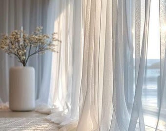 Pode incluir: Cortinas brancas transparentes que vão até o chão pendem em suaves dobras verticais diante de uma janela, permitindo que a luz do sol filtre através do tecido translúcido. Um vaso branco cilíndrico alto contendo delicados ramos florais brancos.
