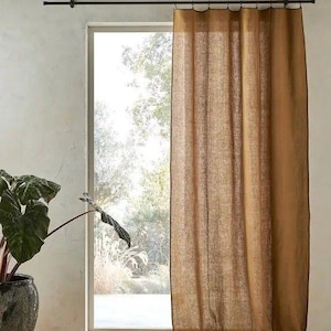 Może przedstawiać: Zasłona z lnianego materiału w kolorze złoto-brązowym wisi na czarnym drążku, obramowując okno z widokiem na zieleń. Widoczna jest faktura zasłony, a po lewej stronie stoi roślina doniczkowa z dużymi, ciemnozielonymi liśćmi.