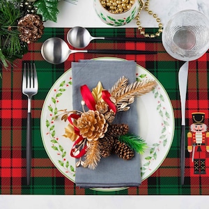 Op de afbeelding: Een feestelijke kersttafelsetting met een rood-groene geruite placemat. Een wit bord met een hulstbessenontwerp bevat een grijze servet en feestelijke decoraties. Bestek en een notenkraker maken de scène compleet.
