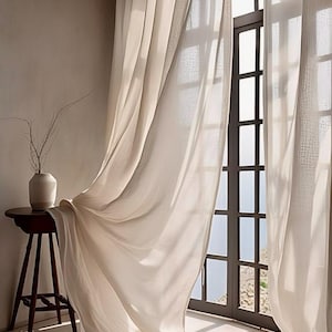 May include: Floor-length, off-white sheer curtains hang in soft folds beside a tall, dark-framed window. A small, round wooden side table holds a simple, light-colored ceramic vase containing thin, bare branches, creating a serene, light-filled scene.