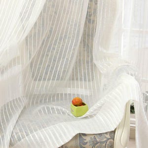 Puede incluir: Una cortina blanca traslúcida con un patrón de rayas verticales de tela semitransparente. El material ligero se extiende sobre una silla, ocultando parcialmente un pequeño recipiente cuadrado verde que contiene una sola fruta naranja redonda.
