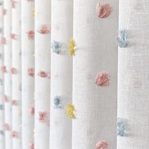 May include: A white fabric curtain featuring a repeating pattern of small, tufted pom-poms in soft pastel shades of pink, light blue, and yellow. The material has a visible woven texture, and the curtain hangs in vertical, pleated folds.