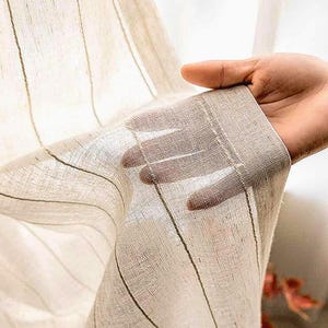 Puede incluir: Un panel de cortina beige claro y transparente con un sutil patrón de rayas verticales. La tela tiene una textura visible de tejido suelto que permite el paso de la luz, creando un efecto semitransparente al sostenerla frente a la luz.