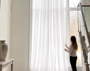 May include: Floor-to-ceiling sheer white curtains covering a large window. The curtains feature vertical pleats and a lightweight, semi-transparent fabric that allows natural light to filter through, creating a soft and airy appearance.