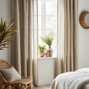 May include: Beige linen curtains frame a bright window, with a wicker chair and a bed with a white duvet in the foreground. A round woven mirror hangs on the wall. Decorative vases and plants sit on the windowsill.