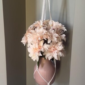 May include: A hanging macrame plant holder with a pink vase filled with light pink flowers. The vase has vertical ridges and is suspended by a white macrame cord from a wooden ring. The flowers are clustered together, creating a full, lush appearance.