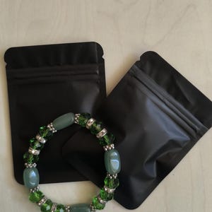 Può includere: Un braccialetto di perline verdi con perline verdi sfaccettate e dettagli argentati. Il braccialetto è esposto con due sacchetti neri su una superficie di colore chiaro. Il braccialetto presenta perline ovali e rotonde.