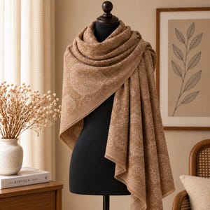 May include: A large, rectangular scarf in a warm tan color featuring an intricate, repeating paisley and floral pattern in a lighter cream shade. The scarf is draped over a dark mannequin, showcasing its soft texture and finished hemmed edges.