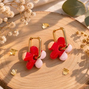 Peut inclure: Une paire de boucles d'oreilles en forme de fleur avec des pétales rouges et roses, et des créoles rectangulaires dorées. Les boucles d'oreilles sont présentées sur une surface en bois avec des accents de feuilles d'or.