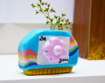 May include: A small, hand-painted ceramic vase in a unique shape, featuring a light blue body with a pink flower and the words "Be you." The vase has a rainbow design on the sides and holds a small sprig of greenery.