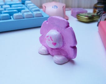 May include: A handmade, pink and white figurine of a cartoon character with a heart on its cheek. The figurine has a fluffy, cloud-like body and is resting on two white, oval-shaped feet. The figurine is on a white surface.