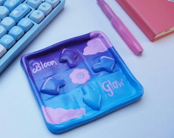 May include: A square, hand-painted trinket tray in shades of blue and purple. It has raised cloud and flower shapes, with the words "Bloom" and "Glow" in pink script. A pink pen, keyboard, and pink notebook are in the background.