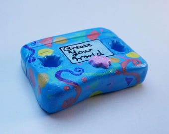 May include: A hand-painted, blue, rectangular object with the text "Create Your World." The object has two circular holes and is decorated with colourful swirls, dots, and a small, pink, star-shaped embellishment.