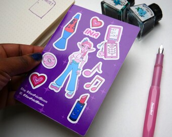 May include: A purple sticker sheet featuring illustrations of a lava lamp, a character, hearts, musical notes, a lipstick, and a flip phone with the text "HEY QT! UDK?". A pink fountain pen and ink bottles are also visible.