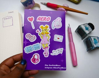 May include: A purple notebook with stickers of a cartoon fairy, hearts, and the word "AERO." The notebook is held near a pink pen, ink bottles, and decorative tape. The notebook has the text "Etsy: SavePointBloom" and "Instagram: @SavePointBloom".