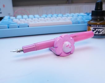May include: A pink fountain pen is displayed on a flower-shaped pen holder. The pen is next to a bottle of brown ink and a bottle of blue ink. A light blue keyboard is in the background.