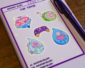 May include: A selection of Y2K-themed stickers on a pale purple surface. The stickers showcase diverse designs, such as a flower, a game controller, and a teardrop shape with text. The text "Y2K TOYS!!" is visible. A purple pen is placed on the side.
