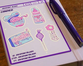 May include: A lilac notebook with a sticker sheet featuring candy-themed illustrations in pink, blue, and purple. The sticker sheet includes text that reads "CANDIES!" and social media handles. A purple pen rests on the notebook.