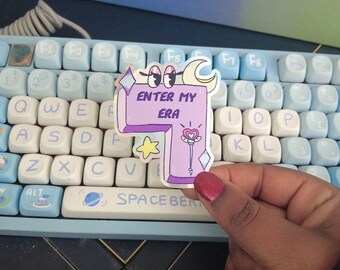May include: A die-cut sticker featuring a light purple, L-shaped design with the text "ENTER MY ERA" in dark purple. The sticker includes cartoon eyes, a yellow crescent moon, a yellow star, and a pink heart-topped wand illustration.