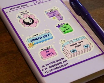 May include: A lilac notebook adorned with vibrant stickers featuring phrases such as "Time to Upgrade" and "Spaced Out." A purple pen lies on the notebook. The text "SAVEPOINT BLOOM" is visible.