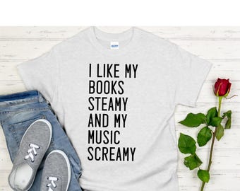 May include: A light gray t-shirt with the text "I LIKE MY BOOKS STEAMY AND MY MUSIC SCREAMY" in black. The shirt is paired with blue jeans and gray sneakers. A red rose is on the right.