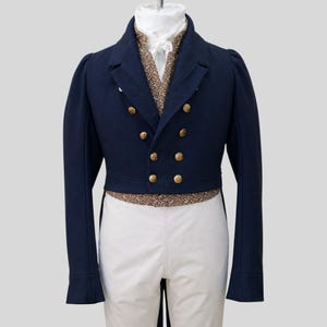 Può includere: Giacca blu navy con bottoni dorati, abbinata a pantaloni bianchi. La giacca ha un design doppiopetto e un colletto alto. Un gilet dorato con paillettes e una cravatta bianca completano l'abito formale. L'outfit è esposto su un manichino.