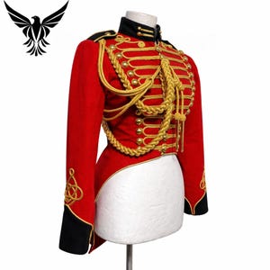 Puede incluir: Una chaqueta roja de estilo militar con trenzas y botones dorados. La chaqueta tiene detalles negros en el cuello y los puños. Una cuerda dorada decorativa cuelga por delante. La chaqueta se exhibe en un maniquí blanco.