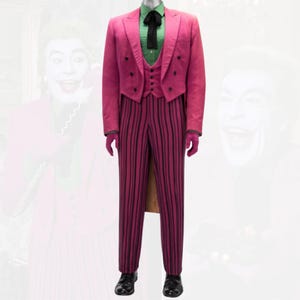 Puede incluir: Un traje rosa con finas rayas negras, compuesto por chaqueta, chaleco y pantalón. La chaqueta tiene un diseño de levita y botones negros. El traje se combina con una camisa verde, una pajarita negra y zapatos negros. Guantes rosas completan el conjunto.