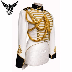 Puede incluir: Una chaqueta blanca de estilo militar con adornos dorados. La chaqueta presenta trenzas doradas, botones y charreteras. Detalles en negro en el cuello y los puños. La chaqueta se exhibe en un maniquí, mostrando su ajuste a medida y sus intrincados detalles.
