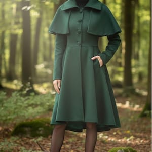 Könnte beinhalten: Ein dunkelgrüner Mantel mit einem kapuzenartigen Kragen und Knopfverschluss. Der Mantel hat eine taillierte Taille und einen ausgestellten Rock. Die Person trägt schwarze Strumpfhosen und Stiefel. Der Hintergrund ist ein Wald.