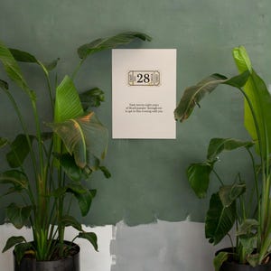 May include: A white poster with the number "28" in a ticket design is mounted on a green wall. The poster includes text that reads "Took twenty-eight years of blood pumpin' through me to get to this evening with you." Potted plants flank the poster.