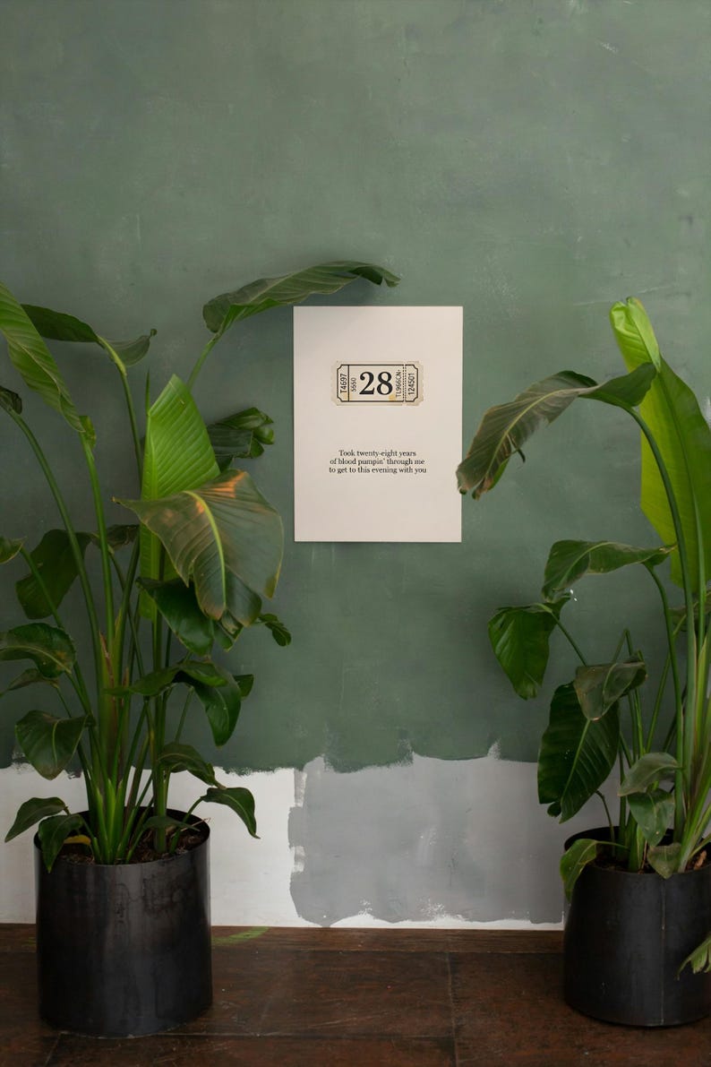 May include: A white poster with the number "28" in a ticket design is mounted on a green wall. The poster includes text that reads "Took twenty-eight years of blood pumpin' through me to get to this evening with you." Potted plants flank the poster.