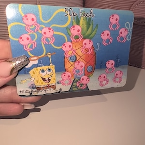 May include: A colourful card featuring SpongeBob SquarePants, a pineapple house, and pink jellyfish in an underwater scene. The text "50p Each" is visible. The card is held against a pink background.
