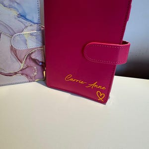 Puede incluir: Dos organizadores de cuero sintético. Uno es de color rosa vibrante con un cierre a juego y el nombre "Carrie-Anne" en escritura dorada, junto con un corazón dorado. El otro tiene un patrón de mármol en tonos rosa, azul y blanco.