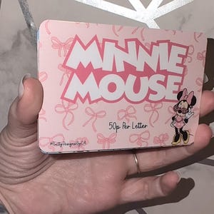 May include: A pink card with the words "MINNIE MOUSE" in white, outlined in pink. The card is decorated with pink bows and a Minnie Mouse illustration. Text includes "50p Per Letter" and "Pretty Designed By CA".
