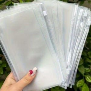 以下が含まれることがあります： 透明なプラスチック製のジッパーポーチの束。各ポーチには白いジッパーと側面に穴の列があります。ポーチは緑の葉の背景の前に保持されています。