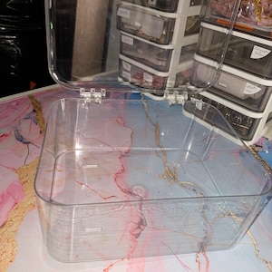 May include: A clear, rectangular plastic storage box with a hinged lid. The container features rounded corners and a transparent design, allowing for full visibility of the interior. The lid is attached with two small, clear plastic hinges.